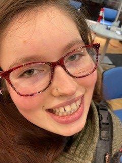 A close-up photo of Lara, a young white woman with long brown hair, brown eyes, glasses and a nose stud smiling up at the camera.